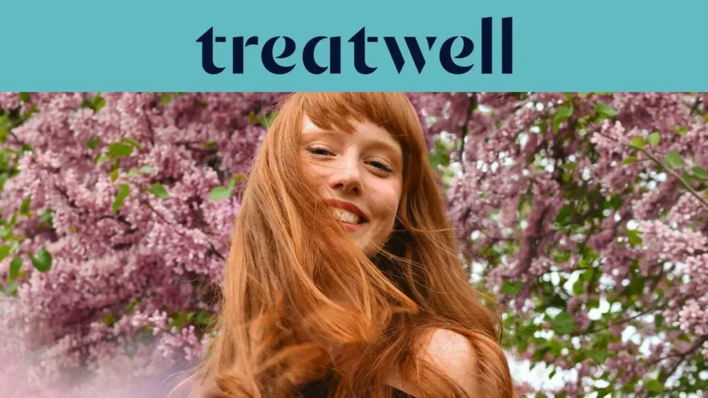 Retrato em primeiro plano de uma jovem ruiva sorridente com franja, com o cabelo a esvoaçar ao vento em frente a um fundo de flores de lilás cor-de-rosa. No topo, uma faixa azul-claro mostra o logótipo da "Treatwell" em azul-escuro.