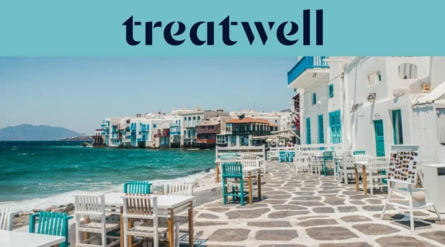 A marginal da Pequena Veneza em Mykonos: casas caiadas de branco com varandas de madeira coloridas à beira da água, o mar Egeu turquesa ao fundo e, em primeiro plano, mesas de café brancas e verde-azuladas dispostas num terraço de pedra.