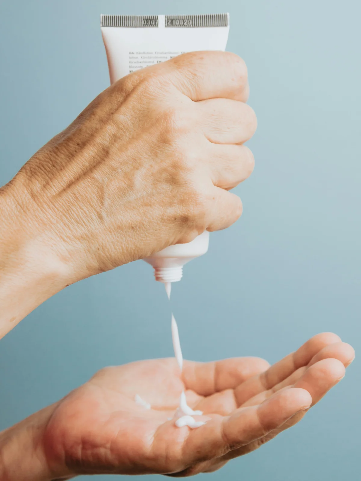 Grande plano de uma mão a segurar na vertical um tubo de creme corporal branco e a deixar cair um pouco na outra mão, por baixo