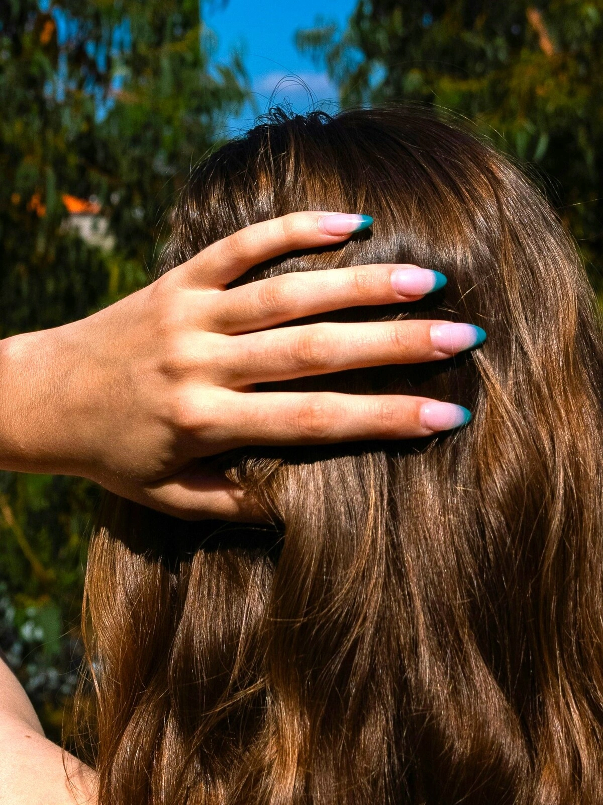 A mão de uma mulher pousada na nuca mostra uma manicure francesa de base rosa-claro e pontas verde-azuladas em unhas amendoadas