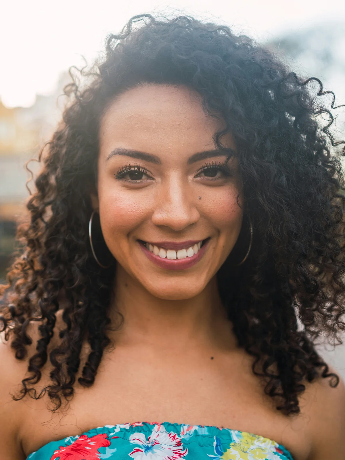 Uma mulher com as sobrancelhas perfeitamente definidas sorri para a câmara, com um top de flores tipo bandeau, ao ar livre com a luz do fim de tarde