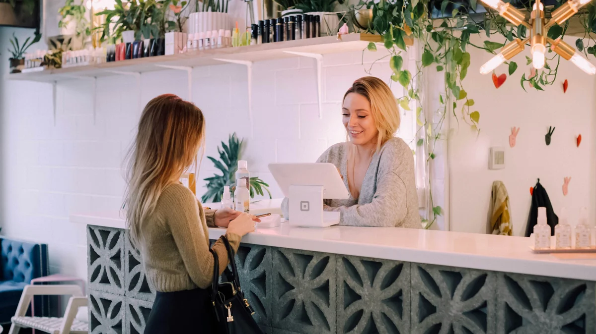 Uma mulher está ao balcão de um salão enquanto uma rececionista marca o seu próximo tratamento; o salão está arrumado, com um balcão branco, prateleiras de produtos bem organizadas e plantas e flores como decoração