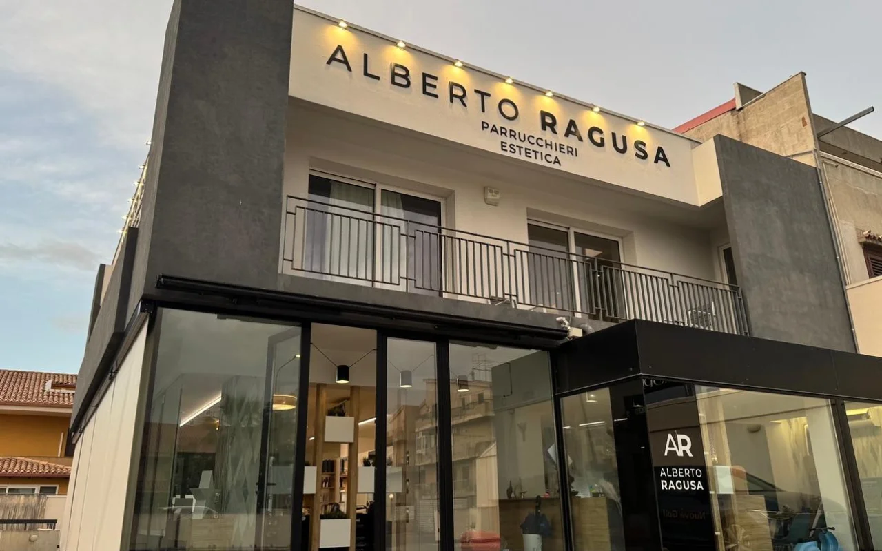 O exterior da Alberto Ragusa ao anoitecer, um salão de dois pisos com um grande letreiro iluminado onde se lê «Alberto Ragusa Parrucchieri Estetica» por cima de janelas do chão ao teto, com uma luz quente a brilhar lá de dentro.