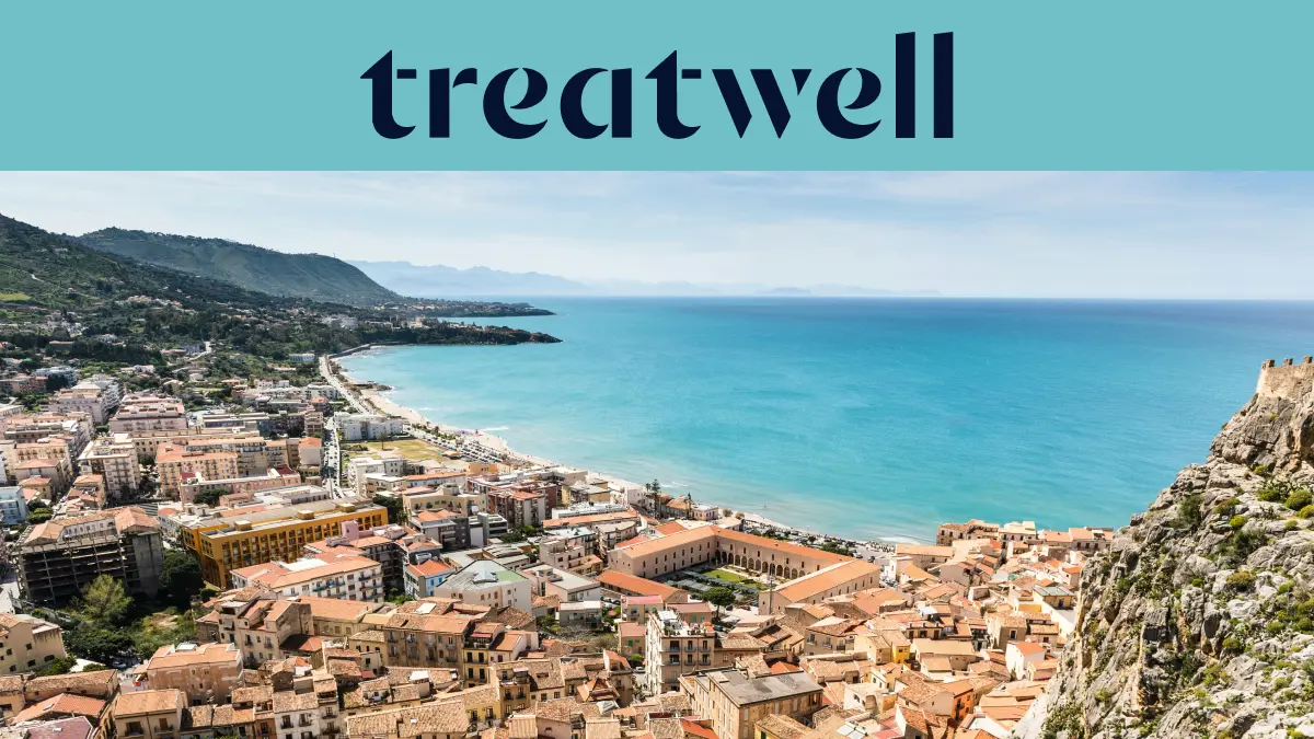 Vista ampla sobre uma cidade costeira siciliana, com edifícios antigos de telhados em terracota, uma meia-lua de praia de areia, um mar turquesa cristalino e um promontório rochoso que se ergue de um dos lados.