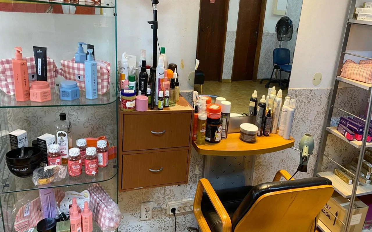 A estação de trabalho de um cabeleireiro com um espelho de parede e uma bancada de madeira repleta de produtos de styling, uma vitrina com produtos de cuidado capilar e suplementos ao lado, e uma clássica cadeira de cabeleireiro em madeira e preto.