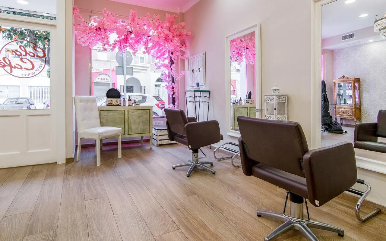 O interior da Color Café Peluqueros em Torremolinos, um cabeleireiro de paredes cor-de-rosa com cadeiras de styling castanhas, grandes espelhos e uma decoração de cerejeira em flor cor-de-rosa na montra.