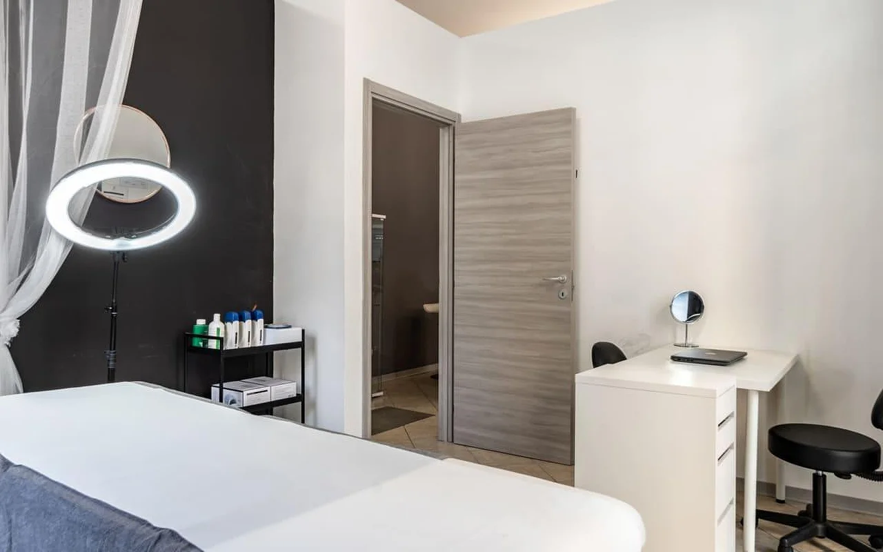 Uma cabine de tratamento na Coquette Wellness and Beauty, com uma marquesa branca, um aro de luz aceso num tripé contra uma parede preta de destaque, cortinados leves na janela e uma pequena secretária branca com um espelho de toucador junto a uma porta que dá para uma casa de banho privativa.