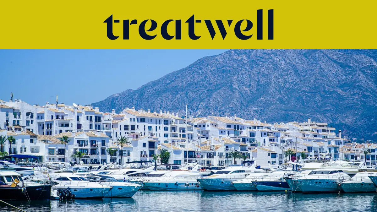 A marina de Puerto Banús, em Marbella, ladeada por edifícios brancos e iates atracados com as montanhas ao fundo, sob um banner amarelo da Treatwell.