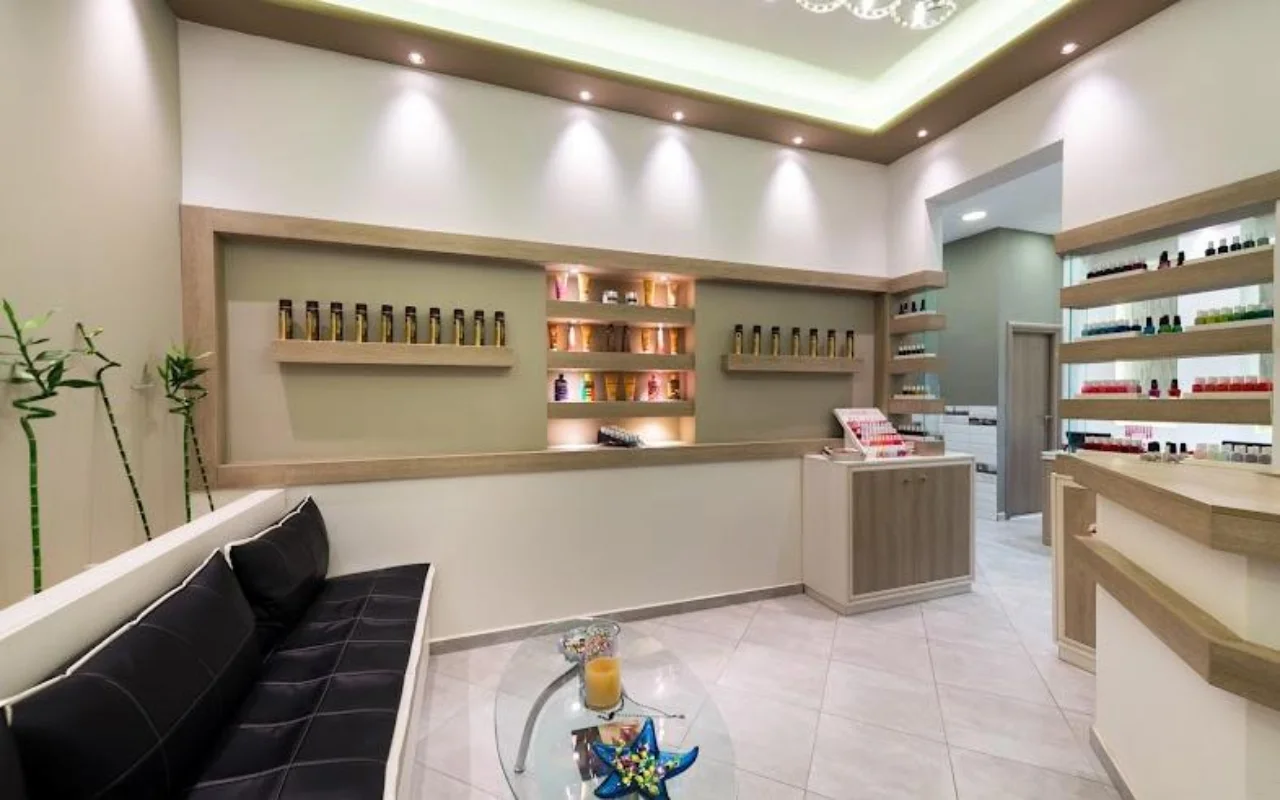 A receção em tons de creme do Fabulous Nails and More em Fira, Santorini, com um banco de pele preta, uma mesa de vidro, prateleiras retroiluminadas com produtos de beleza e luz suave no teto.