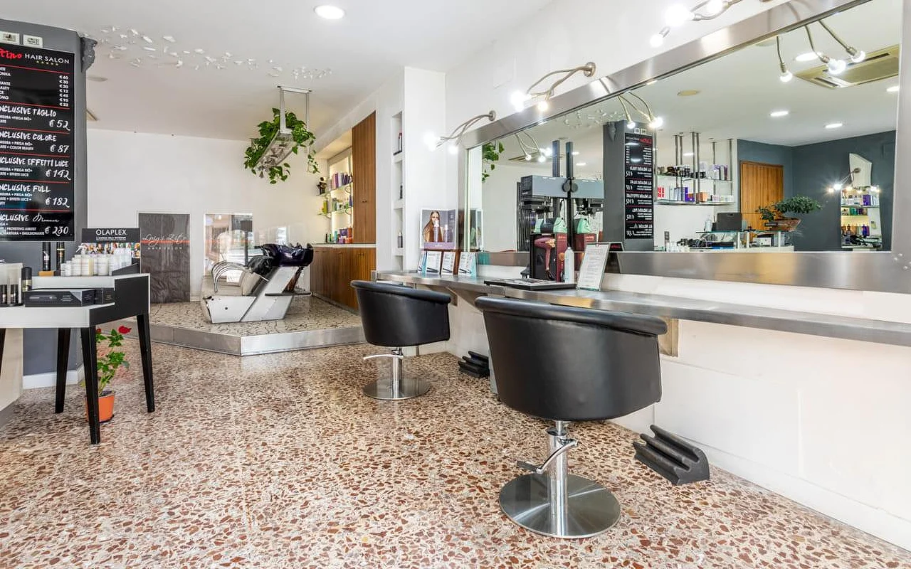 O interior do Hair Salon Luigi Zito, com pavimento em terrazzo, duas cadeiras de penteado pretas em frente a um longo espelho, tabelas de preços em ardósia na parede, um expositor de produtos e um lava-cabeças ao fundo.