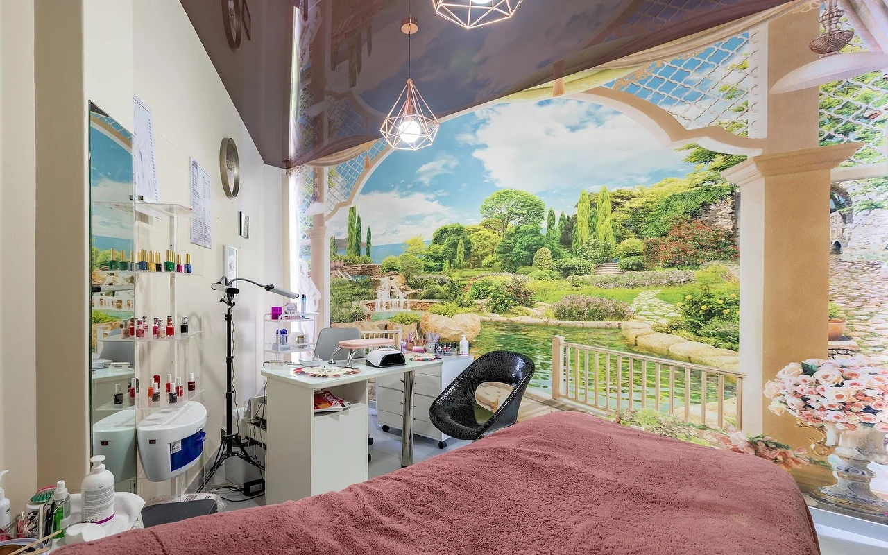 Uma sala de tratamentos de beleza com uma marquesa drapeada de rosa e uma estação de unhas branca, diante de um grande mural de parede com jardim e lago, com prateleiras de vidro abastecidas de produtos ao lado.