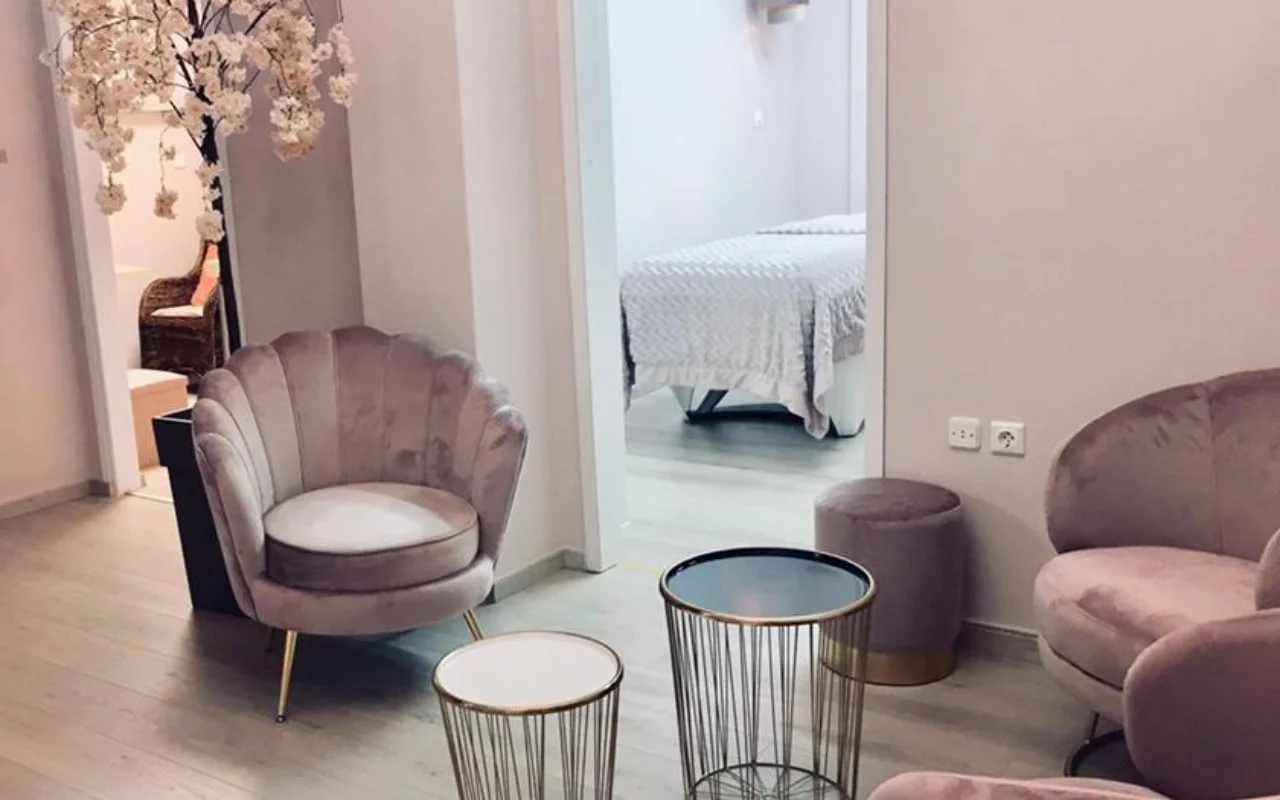 A zona de espera rosa-pó do Nomikou Antonia Aesthetic Lab em Syros, com uma poltrona de veludo de encosto recortado, mesas de apoio douradas finas, um ramo de cerejeira em flor e uma sala de tratamento para lá da porta.