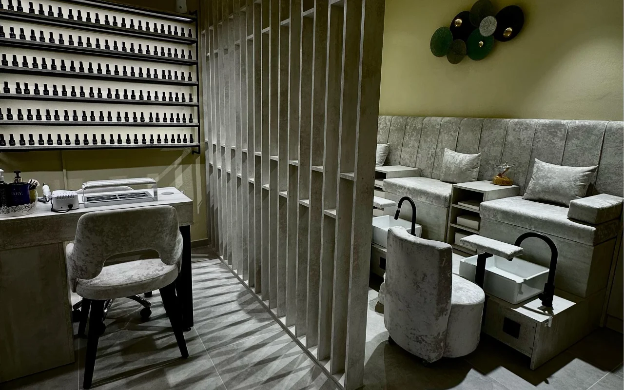 Uma sala de tratamento no Sisu Nail Club and Academy em Perissa, Santorini, com uma parede inteira de vernizes, cadeiras de veludo cinzento para manicure e pedicure e uma divisória de betão perfurado.