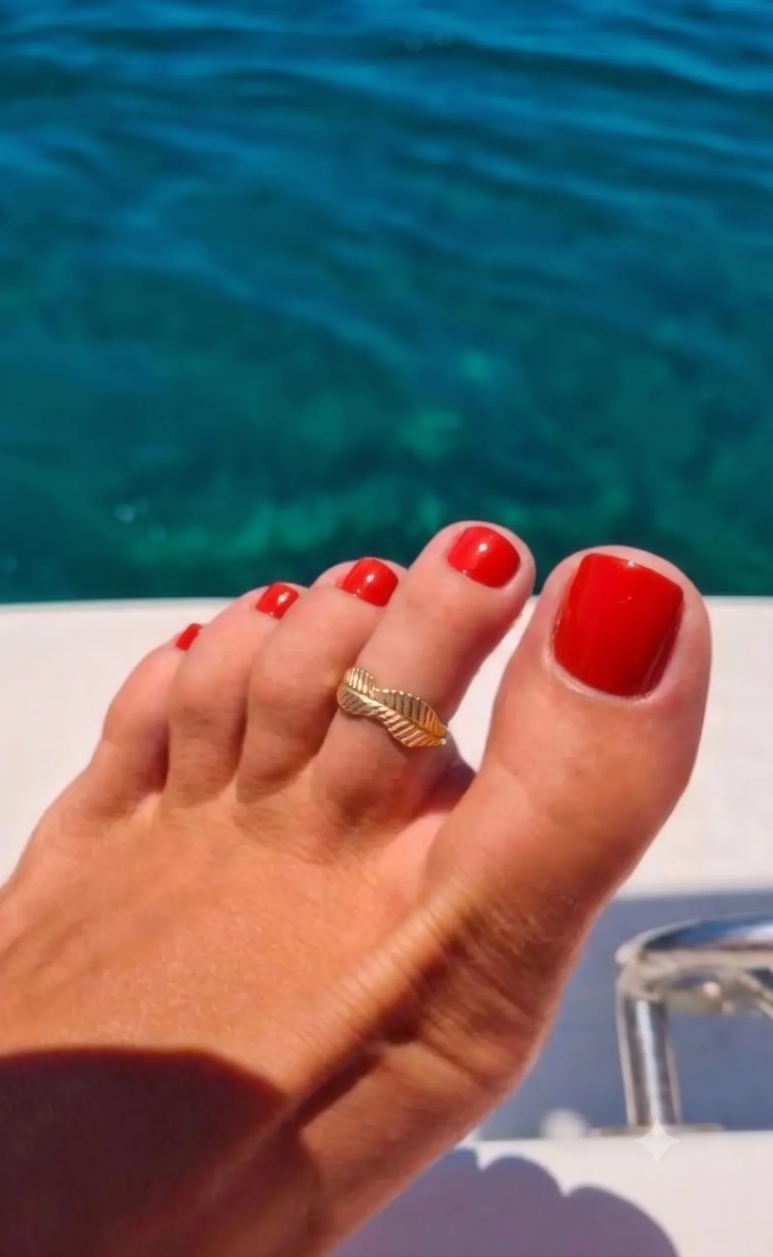 Pedicure em vermelho tomate brilhante num pé bronzeado com um anel de dedo do pé dourado, mar turquesa ao fundo